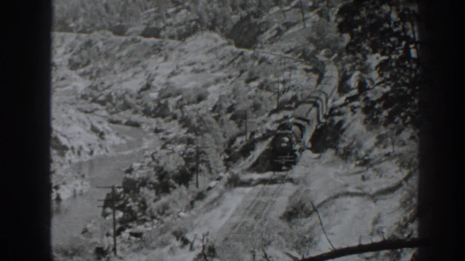 A steam locomotive moves through the Oroville-area canyon rail route, framed by rugged slopes, trees, and the river below.