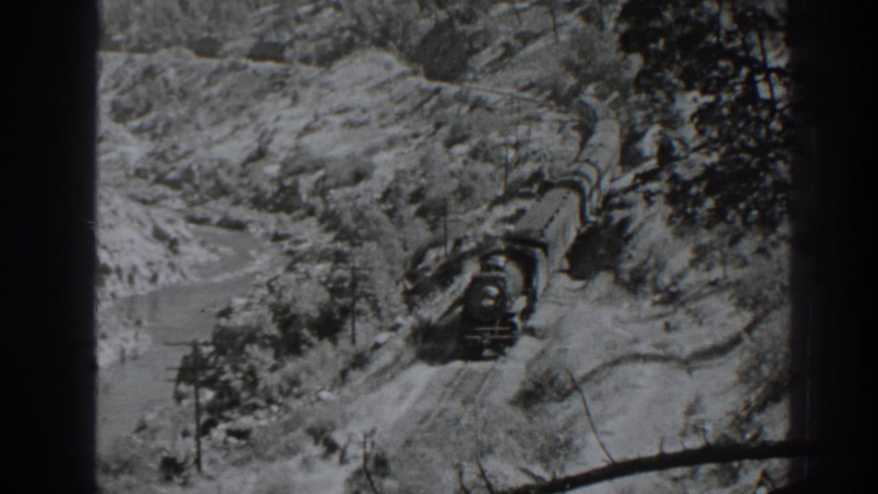 Steam train approaches along snowy mountain tracks beside a river.