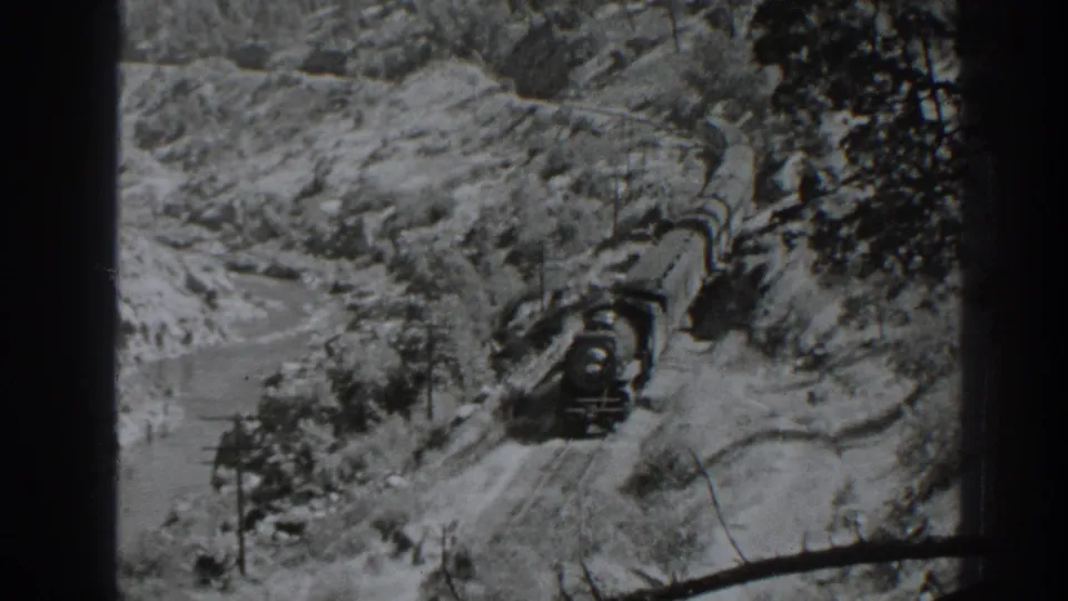 A 1938 Oroville, California 8mm frame shows a steam train moving through a snow-covered mountain pass near water and utility poles.