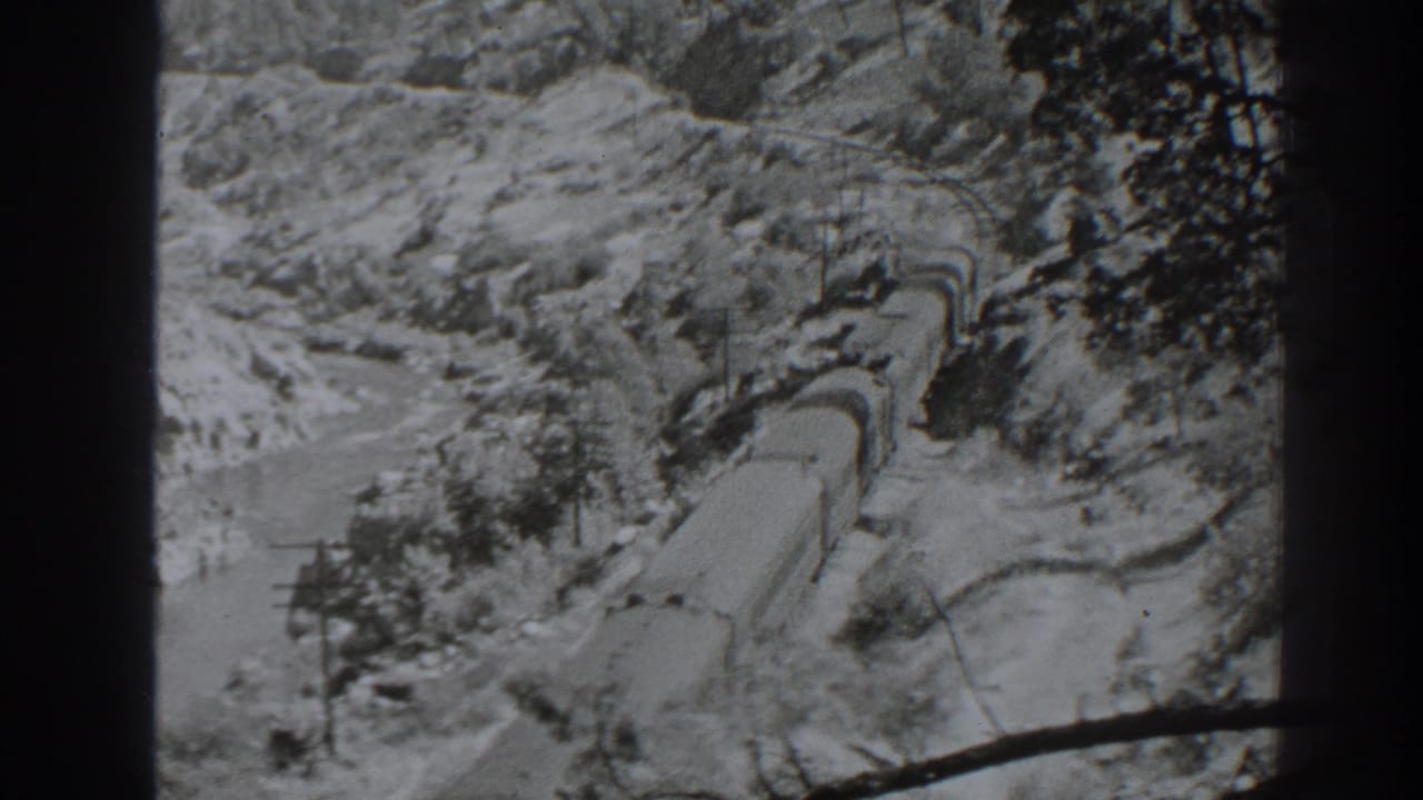 Train cars follow a curve through rocky terrain with trees and a dark film border.