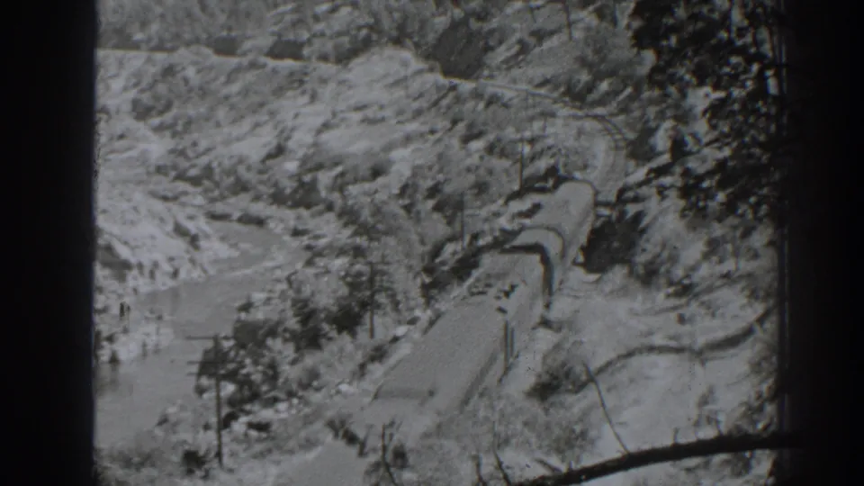 Elevated 1938 Oroville home-movie view of a freight train moving through a rugged corridor with water, poles, trees, and dark frame edges.