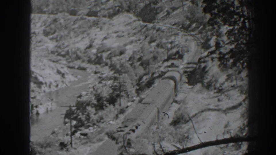 Final 8mm frame shows the freight train in the Oroville mountain corridor, with curving track, rocky slopes, sparse trees, and water below.