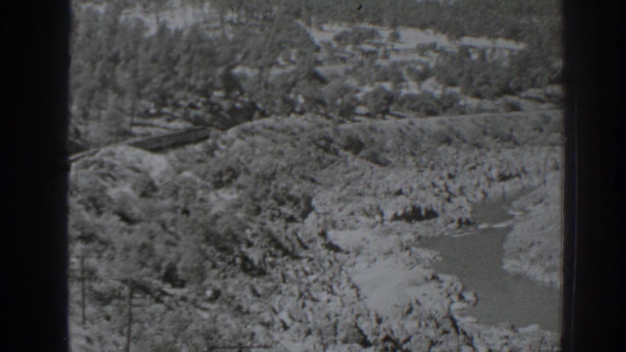 High-angle view of a rocky river bend with wooded slopes and a rail line at left.