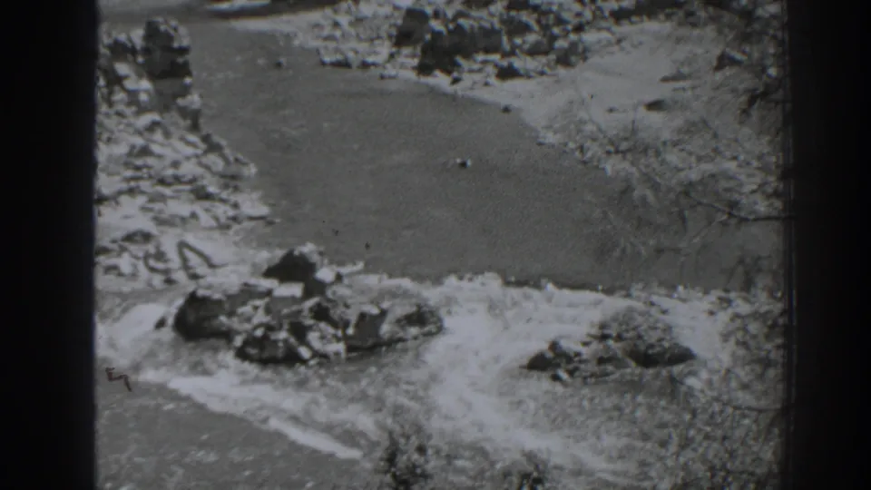 Oroville 1938 home-movie frame showing large rocks in the foreground and a river bordered by pale banks and trees.