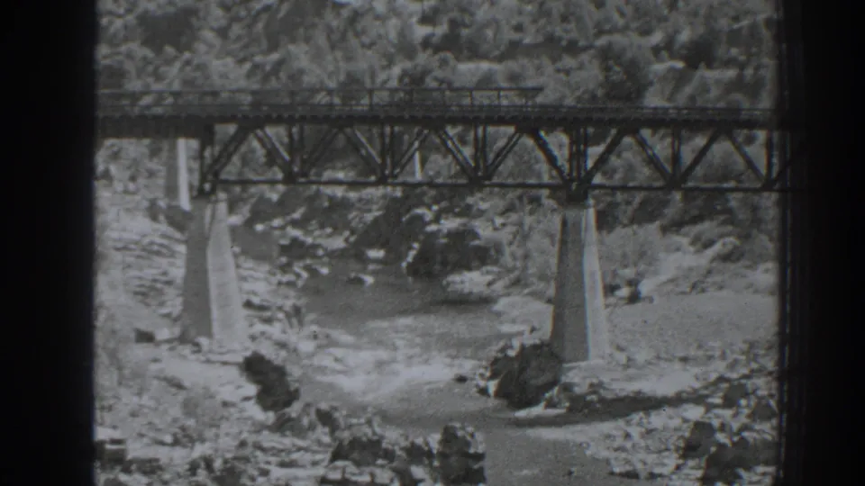 Black-and-white home-movie frame of the Oroville railroad bridge spanning a rocky, fast-moving river.