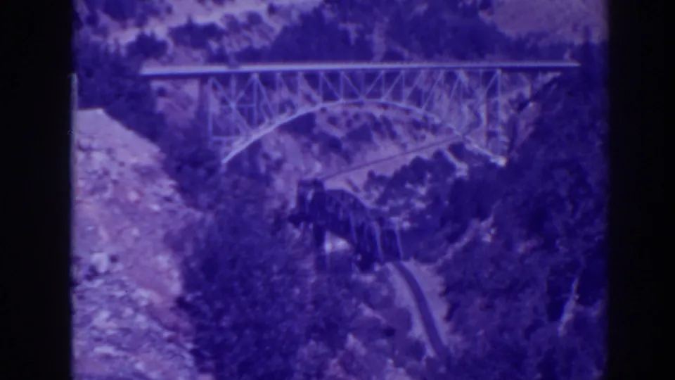 Purple-shifted 1938 Oroville home-movie frame showing the steel bridge, canyon vegetation, and road or path below.
