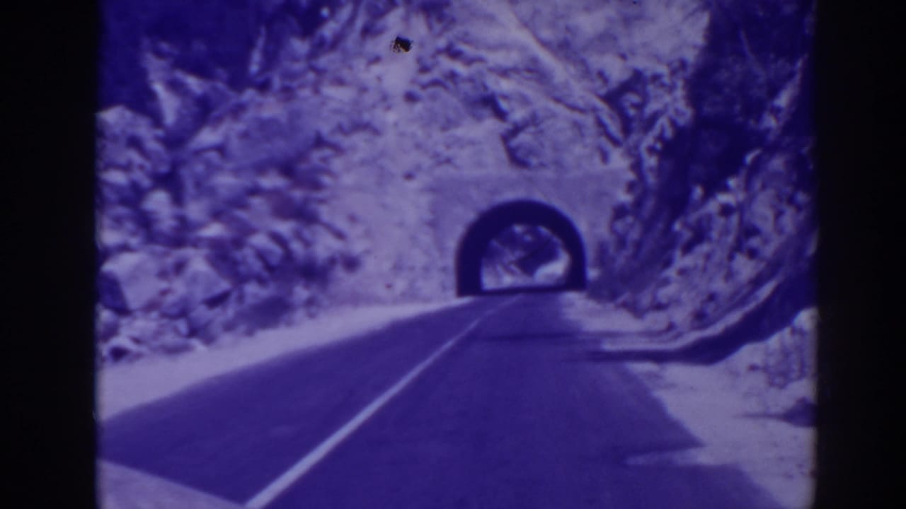 Two-lane mountain road leading toward a dark tunnel in rocky terrain.