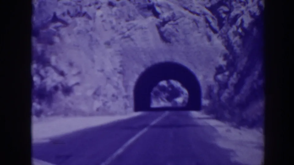 Forward-moving 8mm view in Oroville showing the road closing in on a dark tunnel portal in rocky terrain.