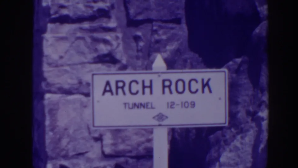 The final frame holds on the Arch Rock Tunnel sign, white post, stone wall, and dark rock edge in aged 8mm color.