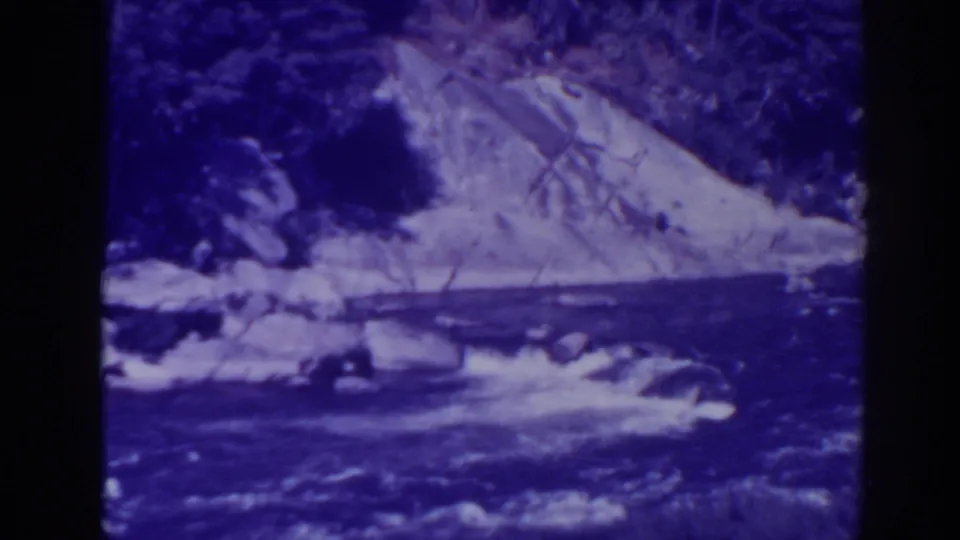 1938 Oroville, California 8mm frame with a rushing stream, scattered rocks, and a pale rocky bank behind it.