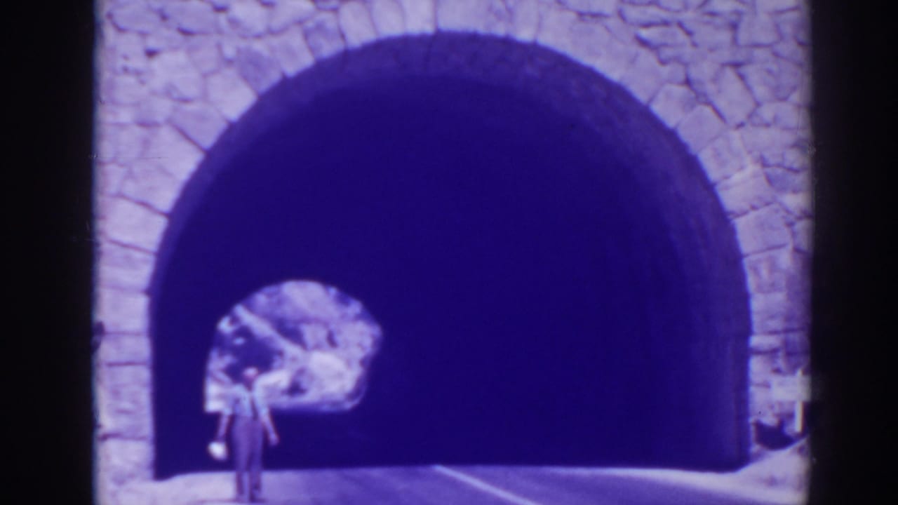 Man standing near a stone-arched tunnel entrance on a mountain road.