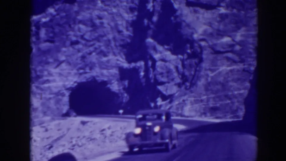 A vintage automobile with headlights on moves along the mountain road near the tunnel entrance in 1938 Oroville footage.