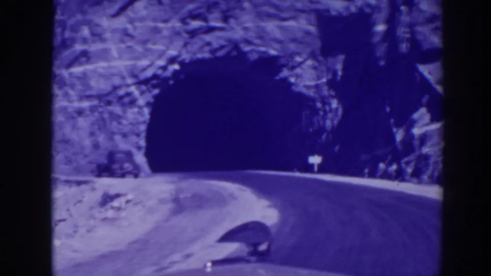 A car continues toward the tunnel on a rocky Oroville roadway, seen in blue-purple 8mm film color.