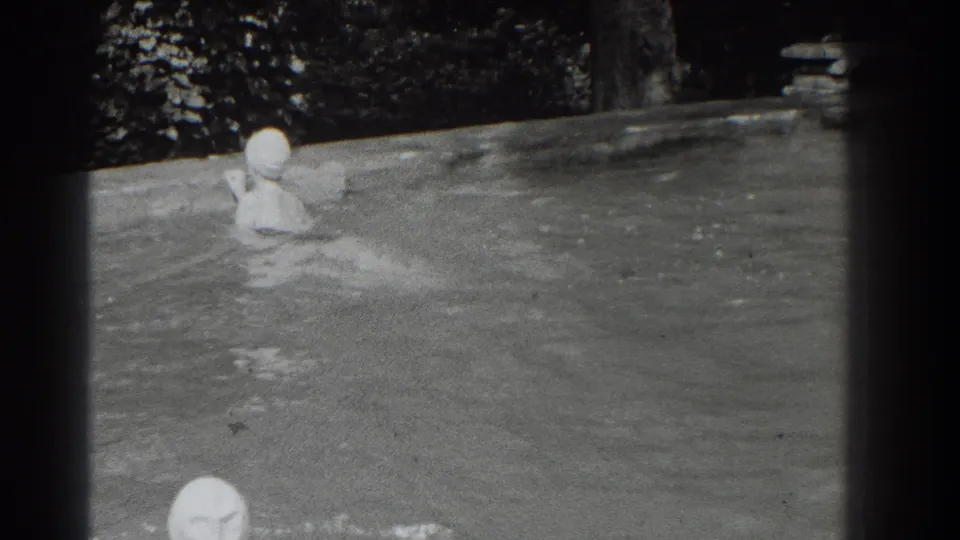 1937 Southampton home-movie frame showing two capped swimmers in a sunlit outdoor pool with trees behind the far edge.