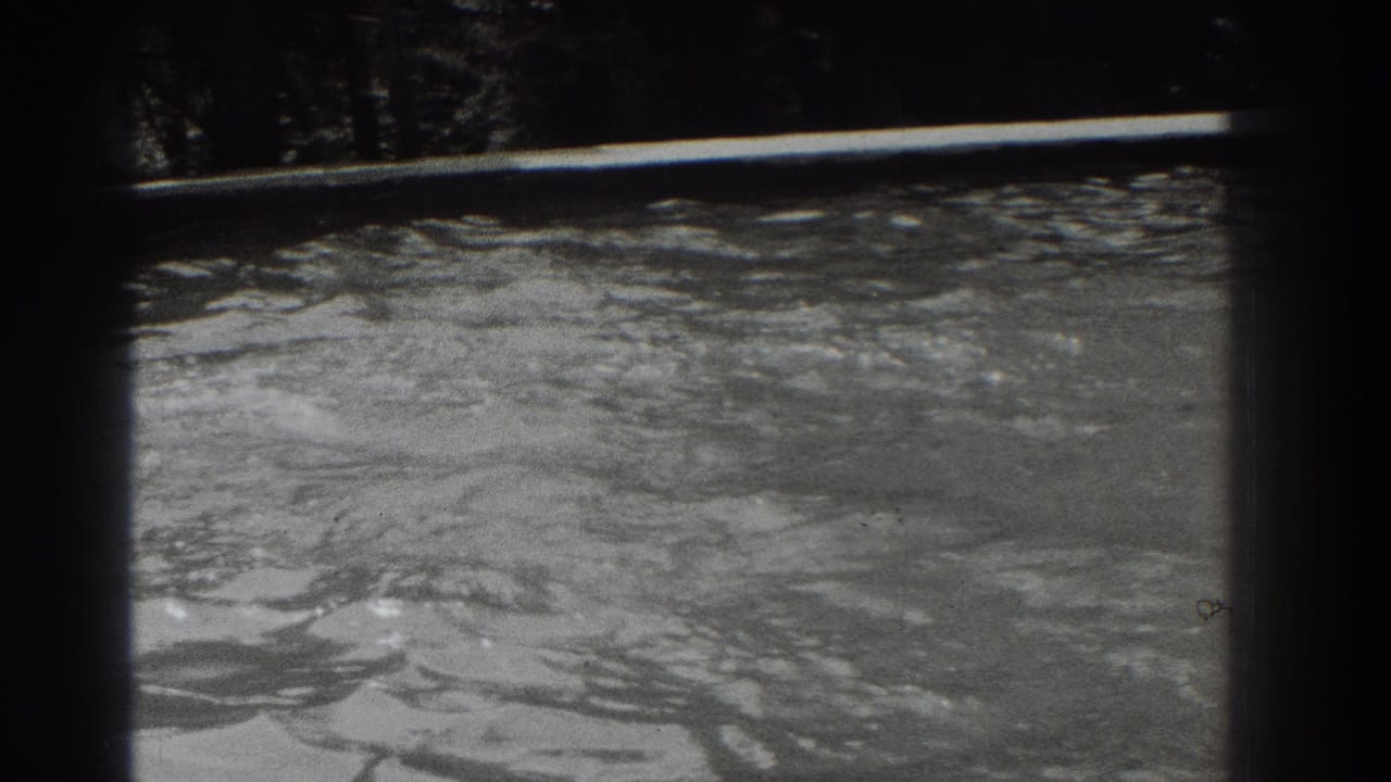 Rippling river water below a dark wooded shoreline in black-and-white 8mm footage.