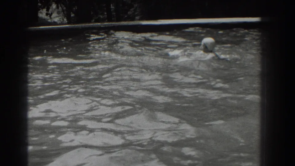 A woman in a white swim cap swims across the outdoor pool, partly submerged amid broad ripples in the black-and-white home movie.