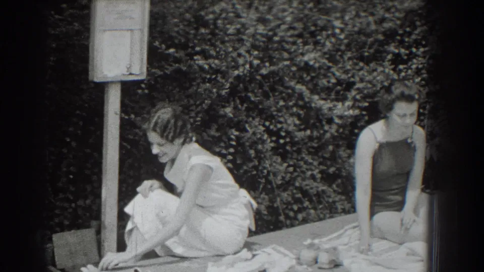1937 black-and-white 8mm frame showing two women seated on poolside towels, one in a light dress and one in a dark swimsuit.