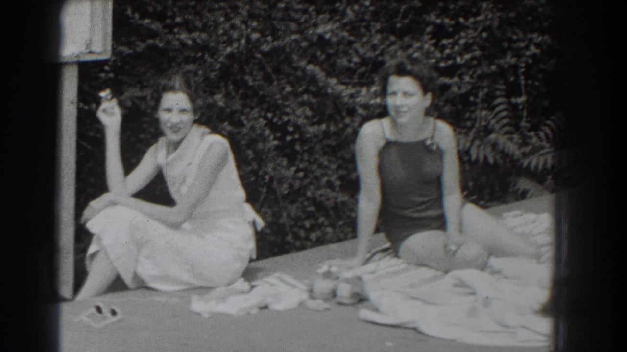 Two women sit on towels outdoors, one holding a cigarette and one in a dark bathing suit.