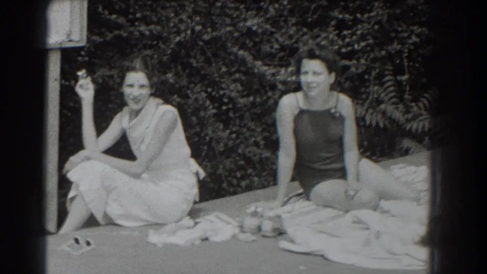 1937 Southampton home-movie frame of two women relaxing on towels near shrubs, with a cigarette and poolside items visible.