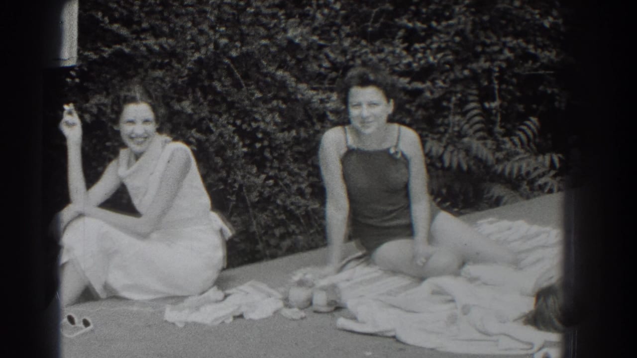 Two women smile on towels beside shrubs, with one holding a cigarette.