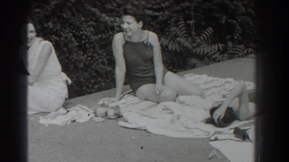 1937 poolside frame with a seated woman in a dark one-piece bathing suit, towels, small items, and a reclining figure at right.