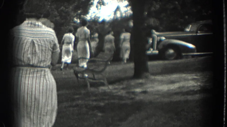 Women in light dresses walk away through a Southampton park, with benches and a period car at the right edge.