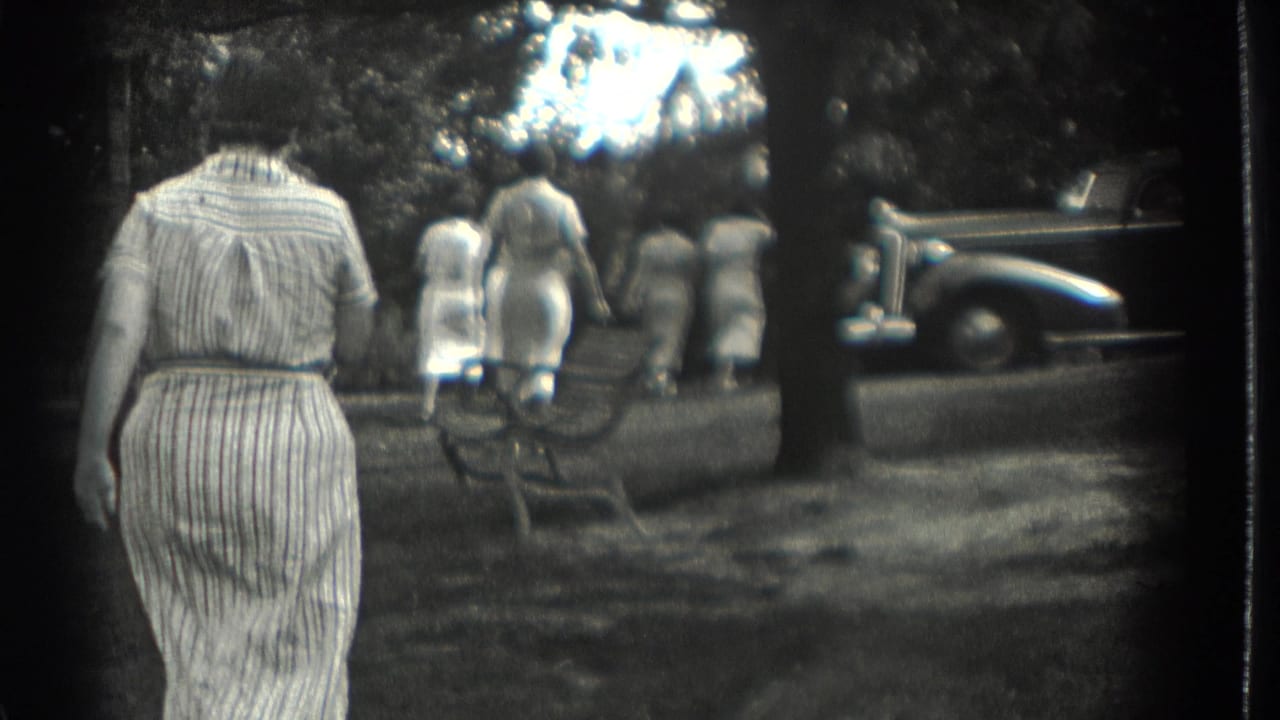 Women in dresses walk through a shaded park near a parked car.