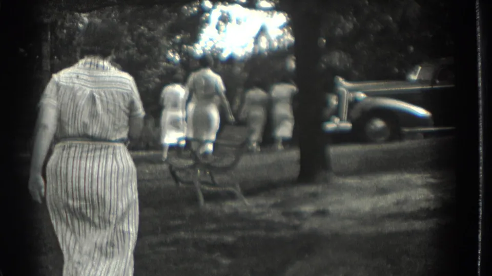 1937 Southampton, New York home-movie frame showing women in dresses walking through a shaded park as a parked car sits at right.