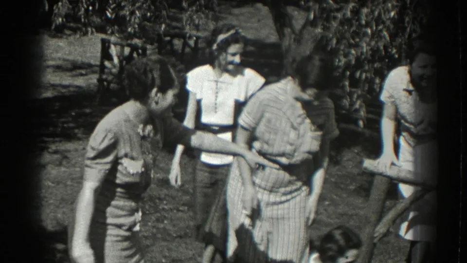 A group in 1930s-style summer clothing stands close together in a Southampton garden, with faces visible and a casual social exchange underway.