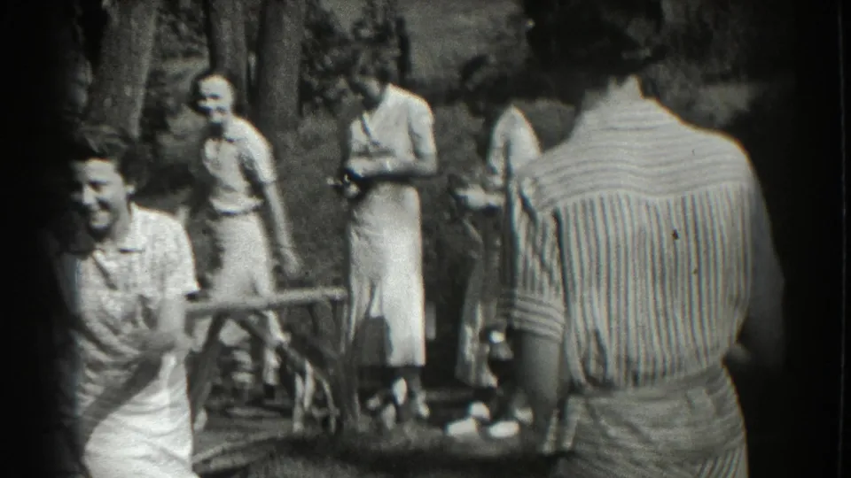 The handheld camera looks across a garden gathering in Southampton, showing smiling adults, dresses, trees, and a figure in a striped shirt from behind.