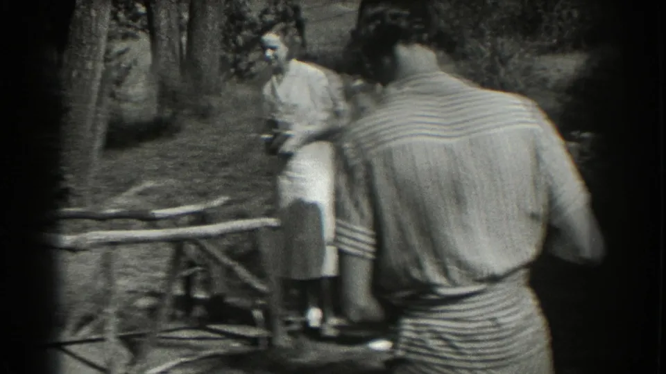 A rear view of a striped-shirted figure frames seated and standing adults in the garden, with black-and-white 8mm softness and partial faces visible.