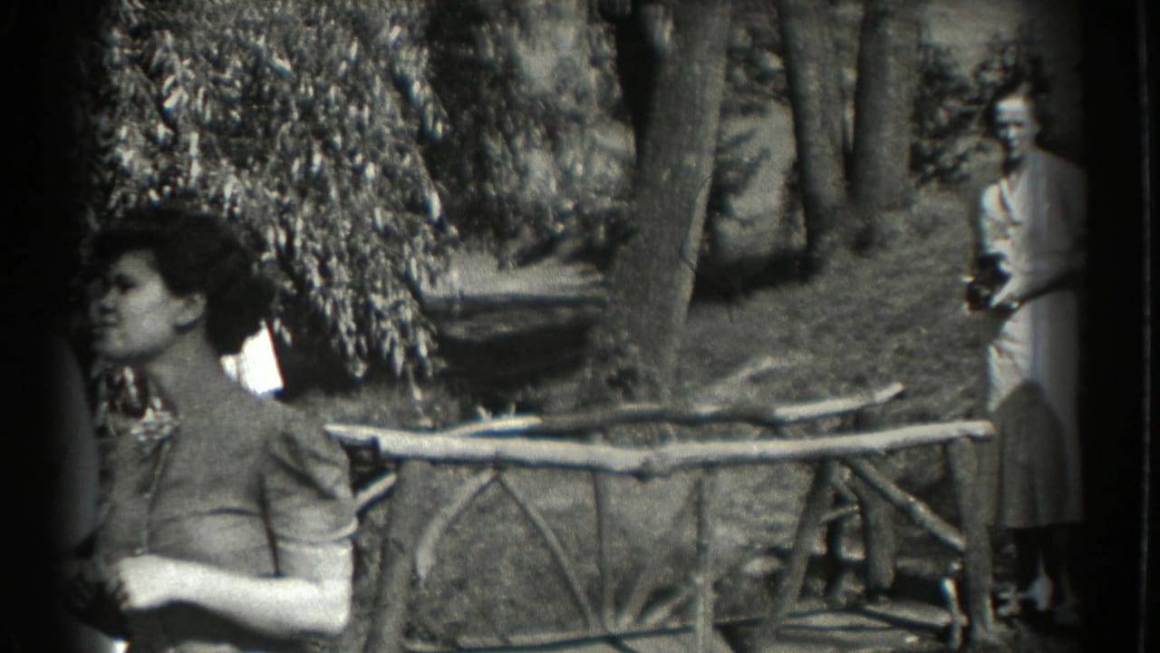Woman near a rustic fence in a wooded garden, with a background figure partly visible.