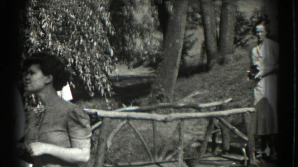 In 1937 Southampton home-movie footage, a woman stands near a rustic branch fence in a wooded garden as another figure appears behind her.