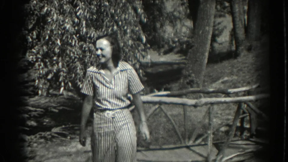 A smiling woman in a striped dress walks through a wooded Southampton garden beside a rustic branch fence in 1937 footage.