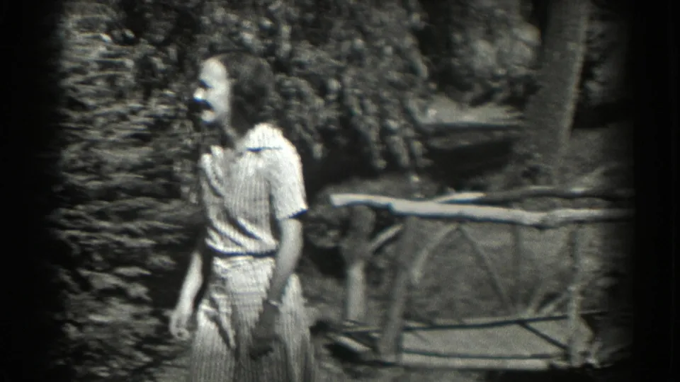 The woman continues walking past leafy shrubs and a rustic fence in the wooded Southampton garden, captured in grainy 8mm film.
