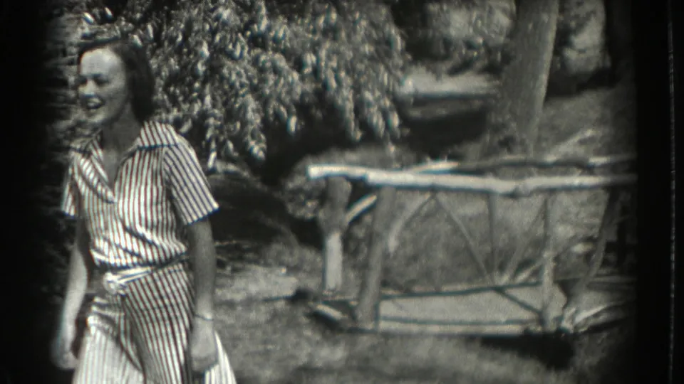 A smiling woman in a striped outfit walks near trees, foliage, and a rustic branch fence in the 1937 Southampton garden scene.
