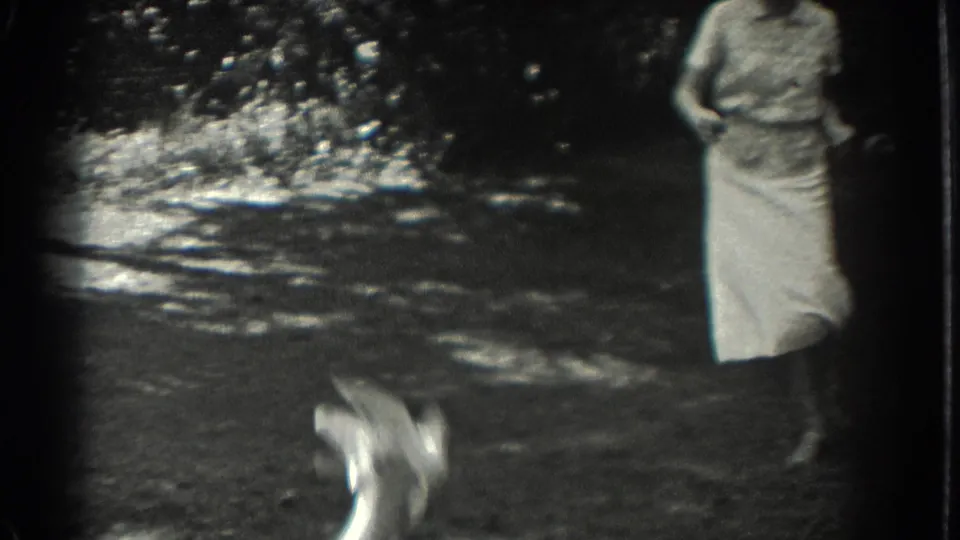 In 1937 Southampton home-movie footage, a woman in a light dress stands in a dappled garden as a blurred figure moves low in front of her.