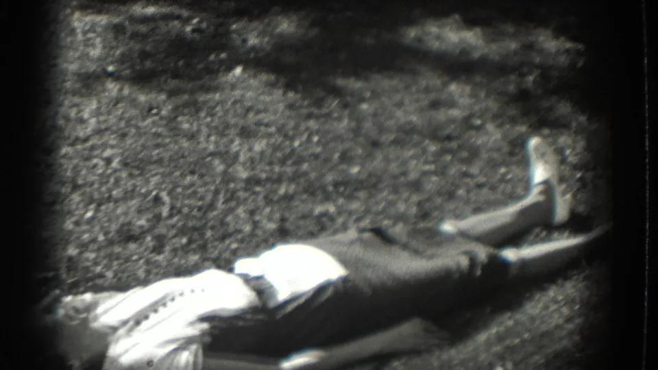 The home-movie frame shows a woman stretched out on the Southampton garden lawn, her light top catching the sun as the background falls into shadow.