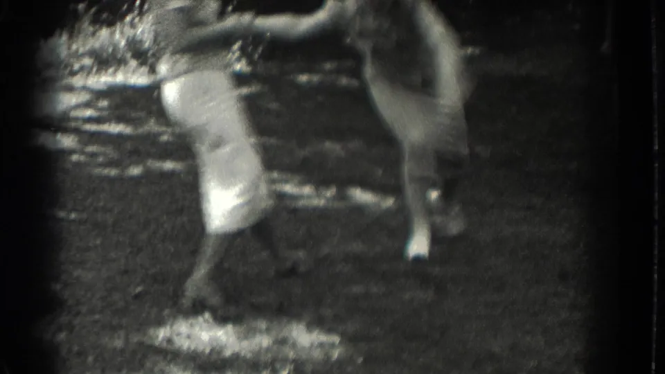 Two women in light dresses are caught mid-motion on the Southampton lawn, one leaning forward as the handheld 8mm camera blurs the action.