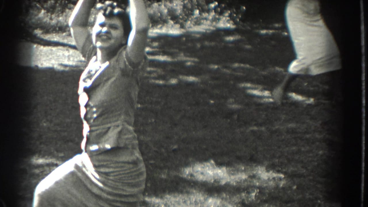1937 Southampton frame of a smiling woman in a dress raising her arms while dancing on a lawn, with a partial onlooker nearby.