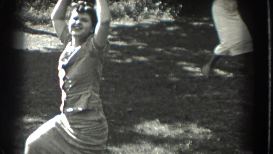 1937 Southampton frame of a smiling woman in a dress raising her arms while dancing on a lawn, with a partial onlooker nearby.