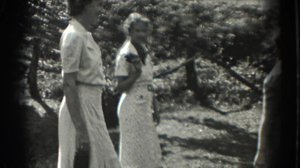 Two adult women in 1930s Southampton stand in a sun-dappled garden, one in trousers and one in a light dress.
