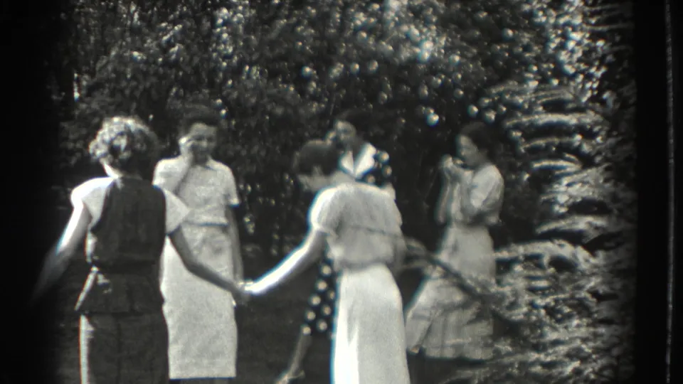 Several adult women in 1930s Southampton gather in a garden and begin holding hands in a loose circle.