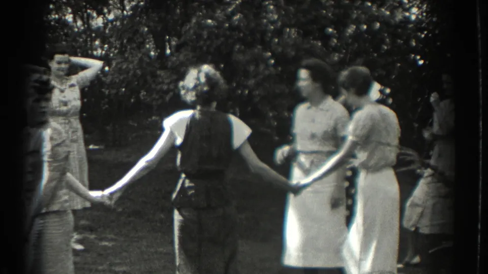 Adult women in summer dresses and trousers hold hands in a Southampton garden circle, with trees and sunlit grass behind them.