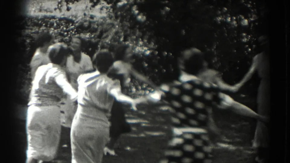 Rear view of women and girls holding hands in a garden ring game, with black-and-white 8mm grain and soft motion.