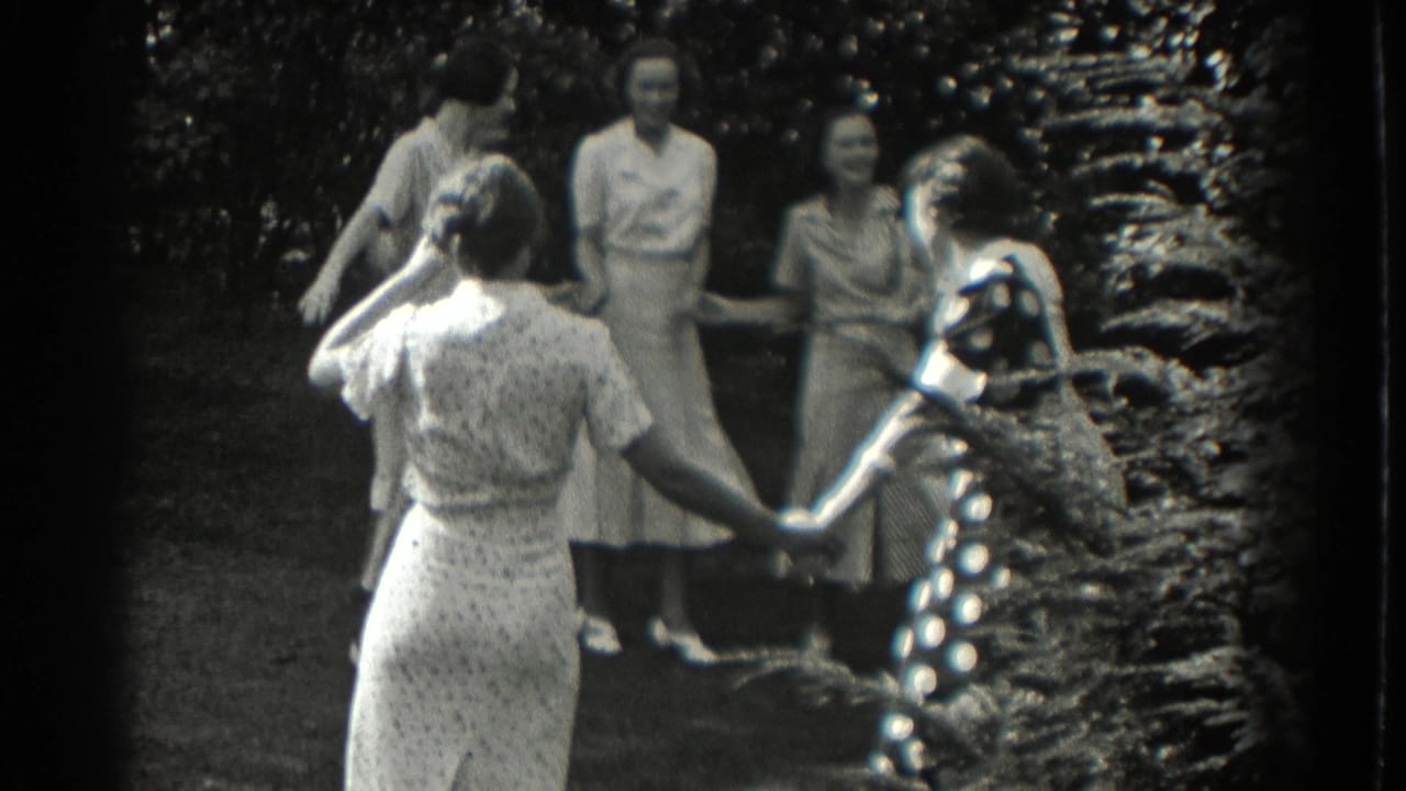 Women in dresses hold hands in a loose circle on a shaded lawn.