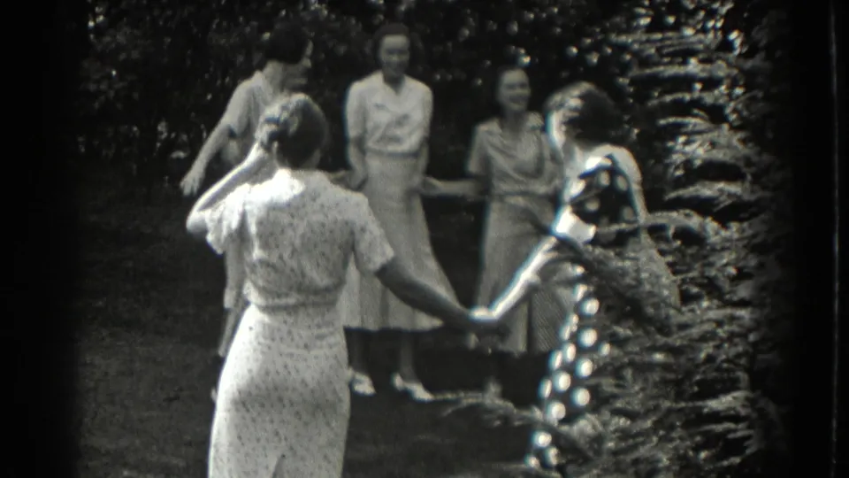 In 1937 Southhampton, New York footage, women in patterned dresses hold hands in a circle on a tree-lined lawn.