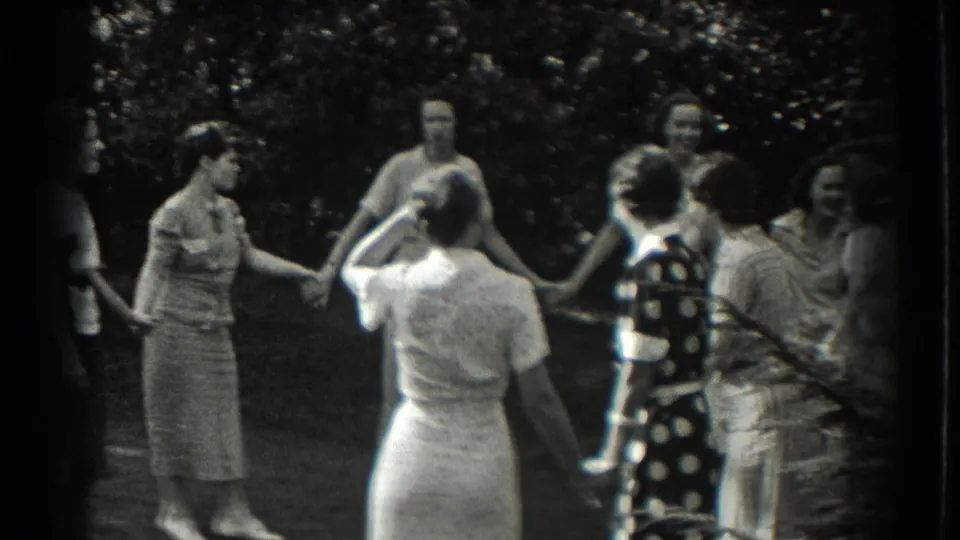 Women in 1930s dresses stand hand in hand in a circle, with trees and lawn behind them in Southhampton, New York.