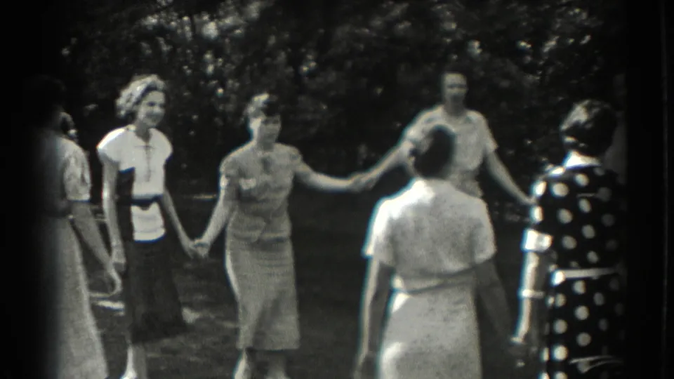 A group of women in period dresses hold hands on a lawn, framed by trees and bright patches of sunlight.