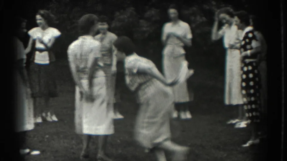 An adult woman runs across the frame while other women in 1930s dresses watch the lawn game in Southampton.